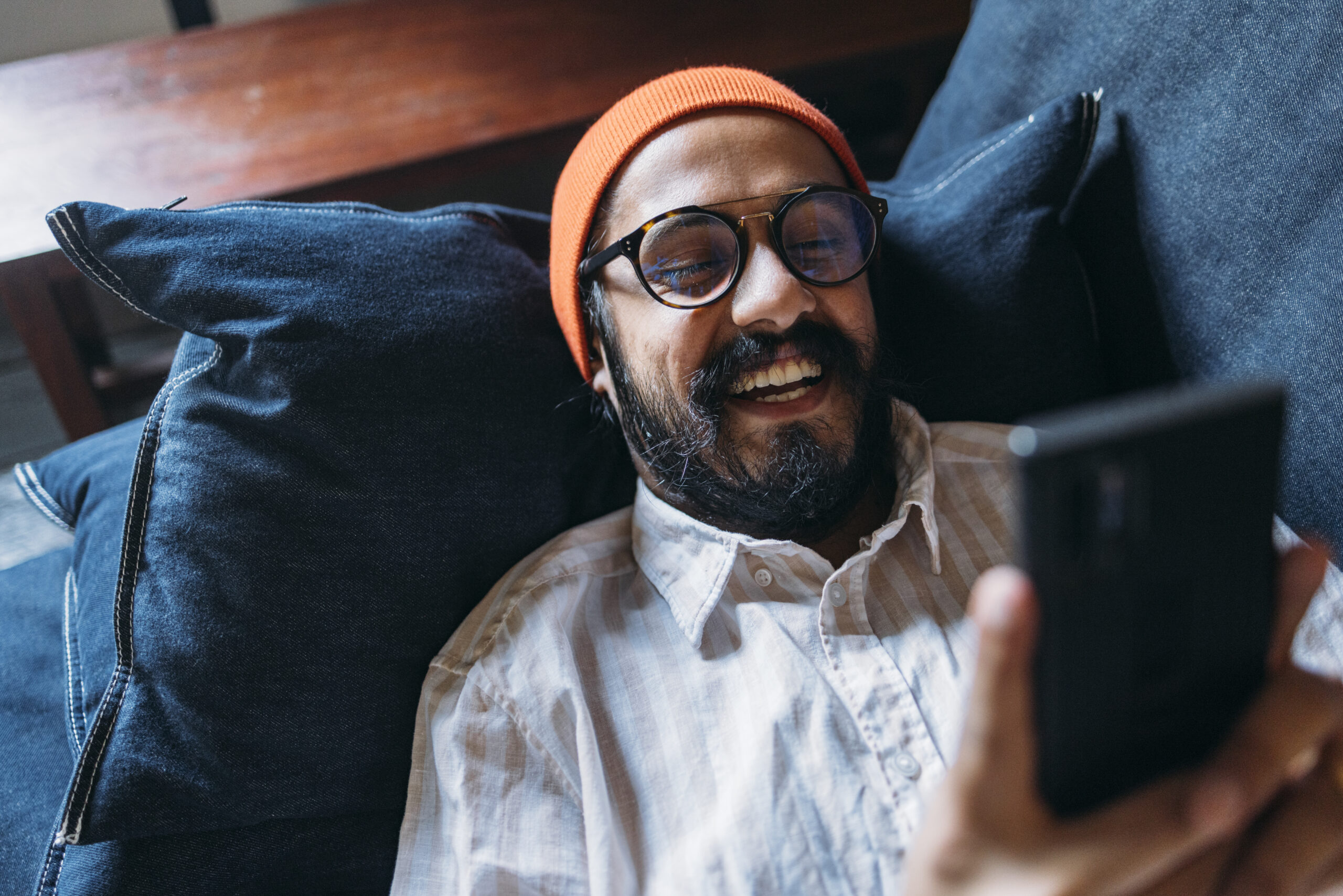 A Happy Handsome Man With Glasses And An Orange Cap On His Head Holding His Mobile Phone While Lying On The Sofa