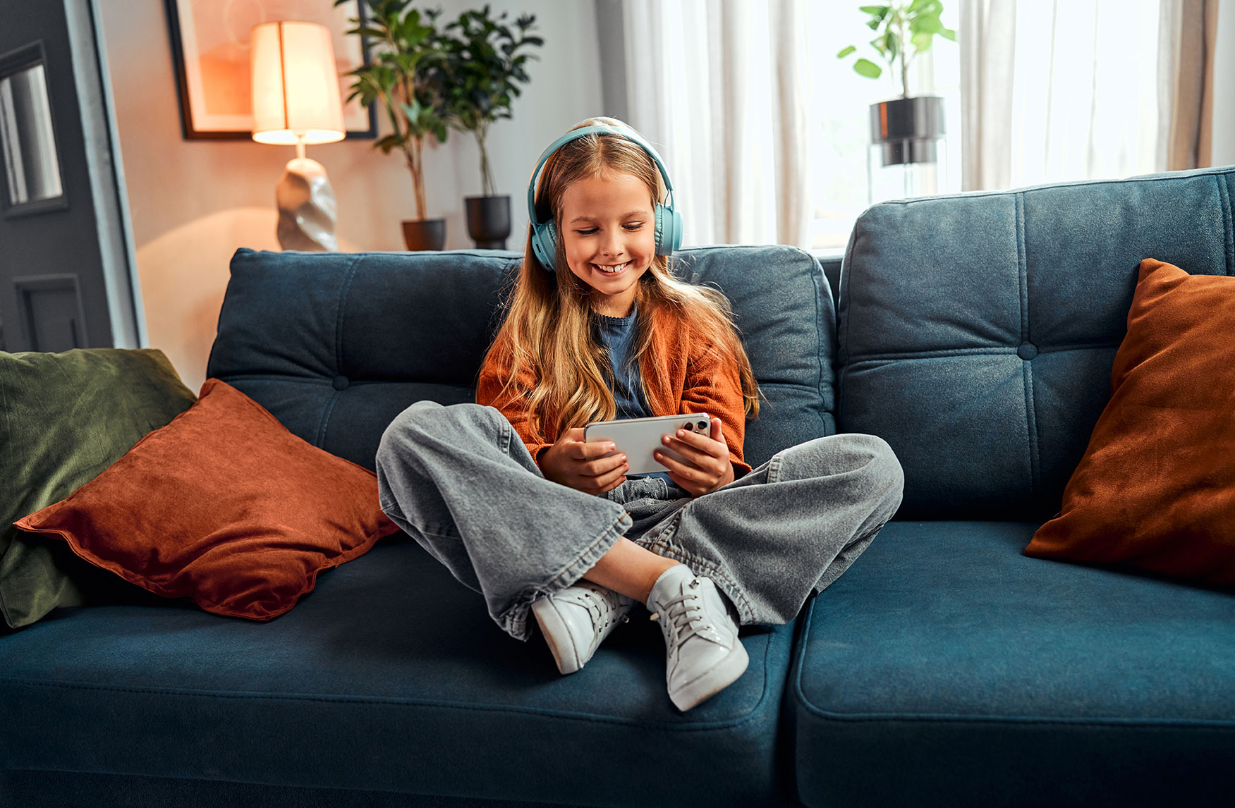 Junges Mädchen sitzt lächelnd auf dem Sofa und spielt auf einem Handy und trägt Kopfhörer.