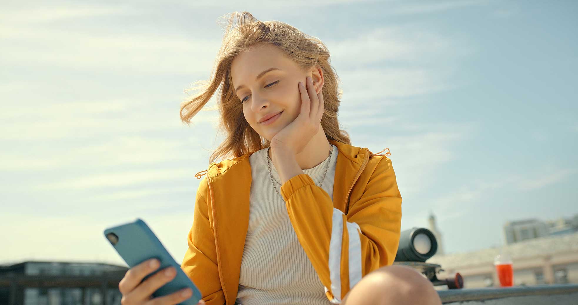 Junge Frau sitzt lächelnd vor einem Handy und geniesst die Sonne.