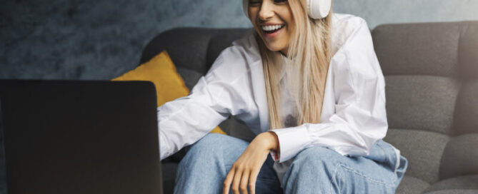 Fröhliches Mädchen mit Kopfhörern sitzt auf dem Sofa vor einem Laptop. Unterhaltungsinhalte für Online-Streaming zu erschwinglichen Abonnementgebühren. Junge Frau schaut sich lustige Videos, Shows und Serien im Internet an.