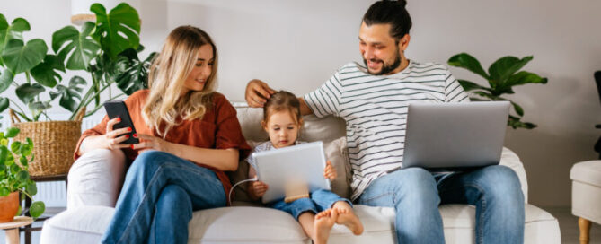 Married couple of different origins and a small preschool daughter are sitting on the sofa, using the computer, watching cartoons online, surfing the Internet, shopping over the Internet and spending the weekend together at home.