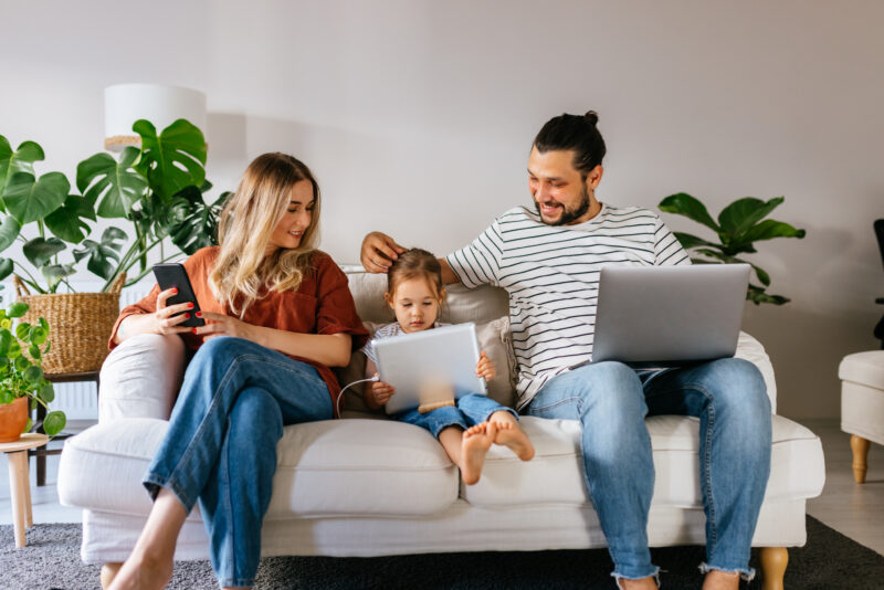 Married couple of different origins and a small preschool daughter are sitting on the sofa, using the computer, watching cartoons online, surfing the Internet, shopping over the Internet and spending the weekend together at home.