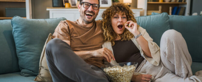 A couple, consisting of a man and a woman of Caucasian origin, husband and wife or boyfriend and girlfriend, sitting at home on the sofa and watching a TV series with a bowl of popcorn, happy smile, connectedness, love, family concept.