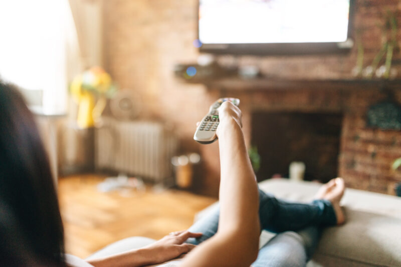 Frau sitzt auf dem Sofa und zappt sich durchs Programm.