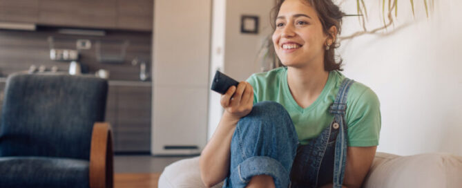 Young woman sits on couch with remote control and channel-surfs.