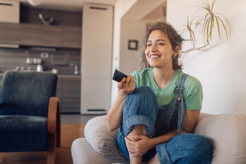 Junge Frau sitzt auf Couch mit Fernbedienung und zappt sich durchs Programm.