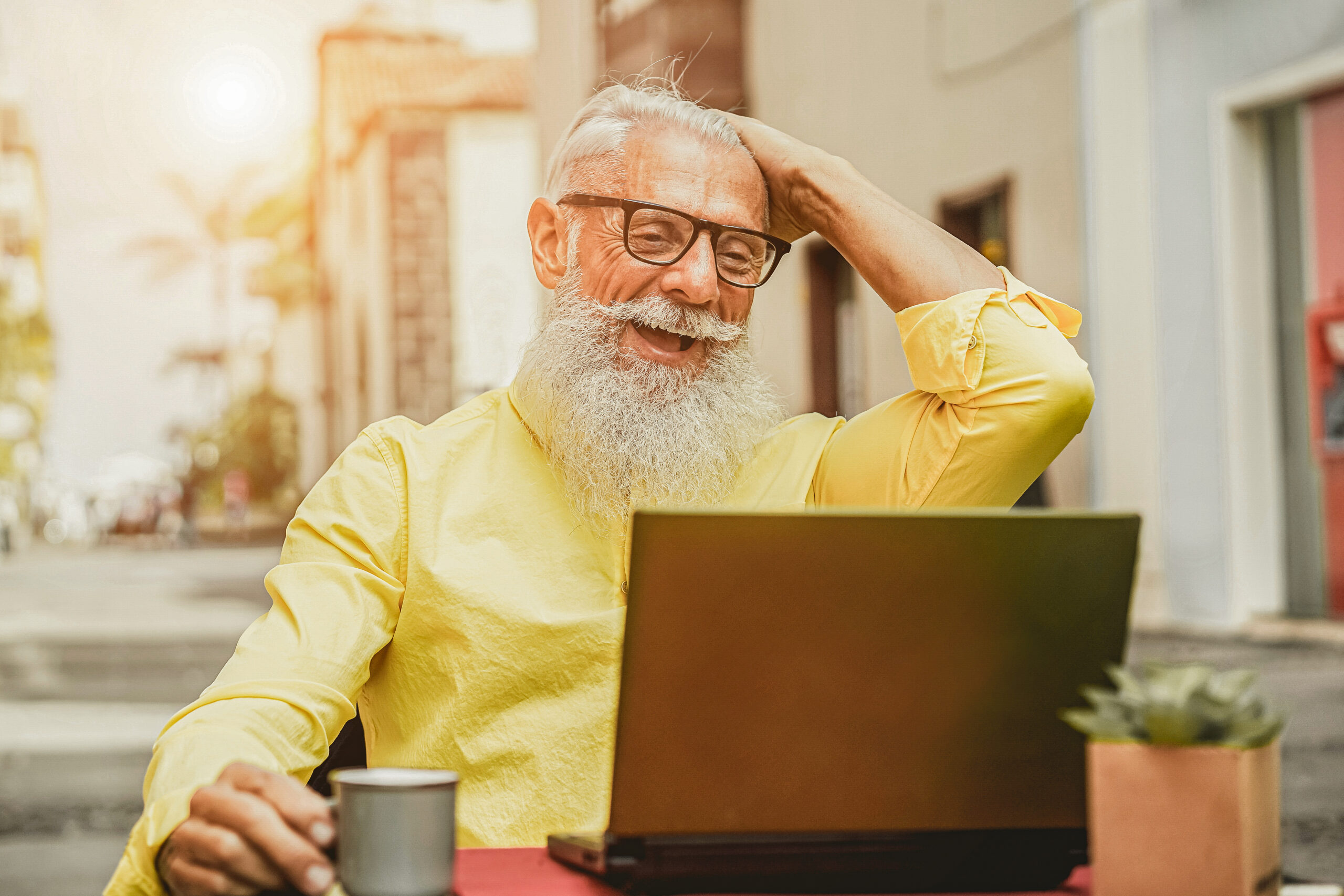Fröhlicher Senior sitzt mit einem Laptop und Kaffee in einem Strassencafé.