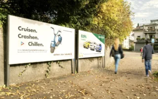 Two advertising spaces at the side of the road and people walking past. 