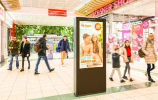 An advertising screen in the middle of the shopping centre with people walking past